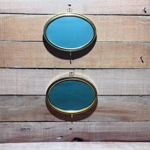 Può includere: Due specchi ovali con cornici dorate e interni color turchese sono montati su una parete di assi di legno. Gli specchi sono sospesi da piccoli elementi decorativi nella parte superiore. Il legno ha un aspetto naturale e invecchiato.