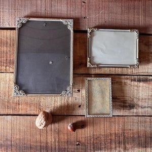 Può includere: Tre cornici antiche con angoli decorati sono disposte su una superficie di legno. Le cornici sono di diverse dimensioni, con una noce e una piccola ghianda nelle vicinanze. Le cornici hanno un'estetica vintage.