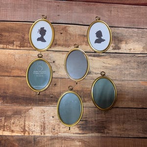 May include: Six oval-shaped antique brass picture frames. Two frames feature black silhouette portraits on a white background. The other four frames have a green or gray background. The frames are displayed on a wooden surface.