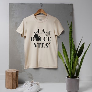 Puede incluir: Una camiseta beige con un estampado gráfico negro que dice "LA DOLCE VITA" con un diseño de limón y naranja.
