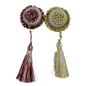 Peut inclure: Deux embrasses de rideaux décoratives. L'une est bordeaux et blanche, l'autre est dorée et blanche. Chacune a un centre rond à motifs floraux, un cordon torsadé et un pompon. Les pompons sont de couleurs assorties.
