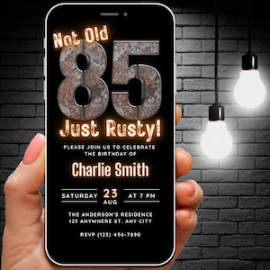 May include: A black smartphone with a birthday invitation on the screen. The invitation is for a 85th birthday party and says "Not Old, Just Rusty!" The invitation is for a party on Saturday, August 23rd at 7 PM. The invitation is for Charlie Smith.