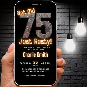 Puede incluir: Un teléfono inteligente negro con una invitación de cumpleaños en la pantalla. La invitación dice "Not Old, 75 Just Rusty!" e incluye detalles para una fiesta celebrando el cumpleaños número 75 de Charlie Smith. El teléfono está sostenido en una mano contra una pared de ladrillo oscuro.