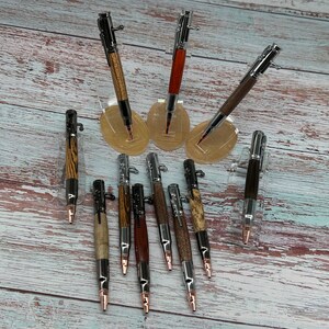 May include: A collection of handcrafted bolt-action pens with various wood grain finishes, including zebrawood, walnut, and maple. The pens have silver and copper-colored metal accents and are displayed on clear acrylic stands against a light wood background.