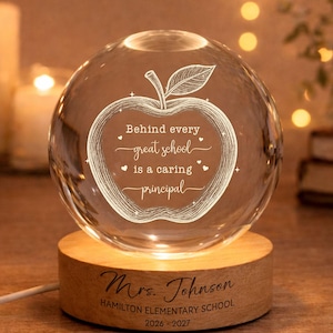 May include: A clear glass sphere lamp on a round wooden base. The sphere features an etched apple design with the text: Behind every great school is a caring principal. The wooden base is engraved with: Mrs. Johnson, HAMILTON ELEMENTARY SCHOOL, 2026-2027.
