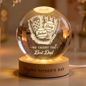 May include: A clear glass sphere with a baseball glove and two baseballs etched inside. The baseballs are inscribed with the names "Brady" and "Jackson." The base is wood and reads "HAPPY FATHER'S DAY." The sphere is lit from within.