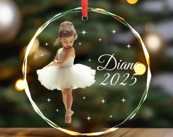 May include: A clear, faceted glass Christmas ornament with a red ribbon. The ornament features a photo of a young girl in a white tutu and tiara, with the name "Diana" and the year "2025" in elegant script.