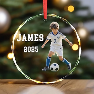 May include: Clear glass ornament with a soccer player image. The ornament has the name "JAMES" and the year "2025" printed in white. A red ribbon is attached for hanging. The soccer player is wearing a white and blue uniform and kicking a soccer ball.