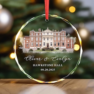 May include: A clear, round glass Christmas ornament with a red ribbon. The ornament features a detailed image of a stately brick building with the names "Oliver & Evelyn", "HAWKSTONE HALL", and the date "08.20.2025" in elegant script.