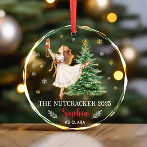 May include: A clear, circular Christmas bauble with a red ribbon. The bauble depicts a ballerina with a nutcracker and a Christmas tree. Text on the bauble reads "THE NUTCRACKER 2025" and "Sophia AS CLARA".