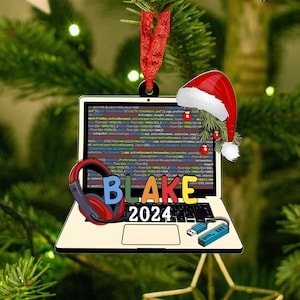May include: A Christmas tree ornament shaped like a laptop with the name "BLAKE" and the year "2024" in colorful letters. The laptop screen displays code, and a Santa hat and headphones are added. A red ribbon is attached for hanging.