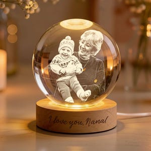 May include: A clear glass sphere with a photo of a baby and an older woman, resting on a wooden base. The base has the inscription "I love you, Nana" engraved on it. The sphere is illuminated from within, creating a warm glow.