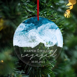 May include: Round clear ornament with a photo of ocean waves crashing on a beach. The text "HARPER & WILLIAM engaged 30.05.2023" is printed on the ornament.