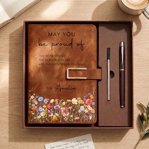 May include: A brown leather-look journal with a gold-tone metal clasp, featuring the printed text 'MAY YOU be proud of THE WORK YOU DO THE PERSON YOU ARE AND THE DIFFERENCE YOU MAKE' and 'FOR Amelia', with a colourful wildflower print and a matching pen.