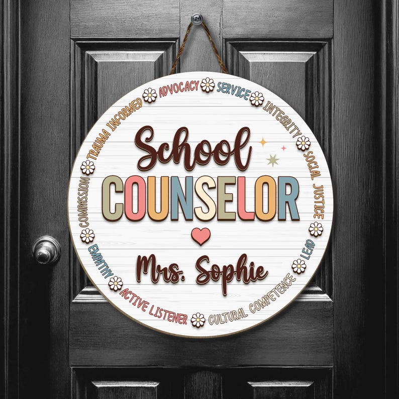 May include: A round wooden sign with the words "School Counselor" in colorful letters. The sign includes the name "Mrs. Sophie" and a heart symbol. The sign is decorated with floral accents and hangs from a brown rope.