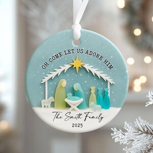 May include: A round Christmas ornament with a light blue background and a white bottom half. The ornament features a nativity scene with the text "OH COME LET US ADORE HIM" and "The Smith Family 2025". A white ribbon is attached.