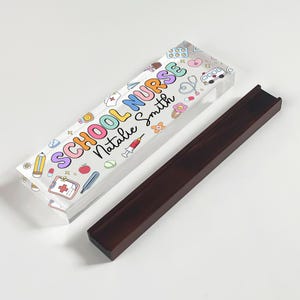 May include: A clear acrylic pen holder with the words "SCHOOL NURSE Natalie Smith" in colorful letters and medical-themed illustrations. Beside it is a dark brown wooden pen holder.