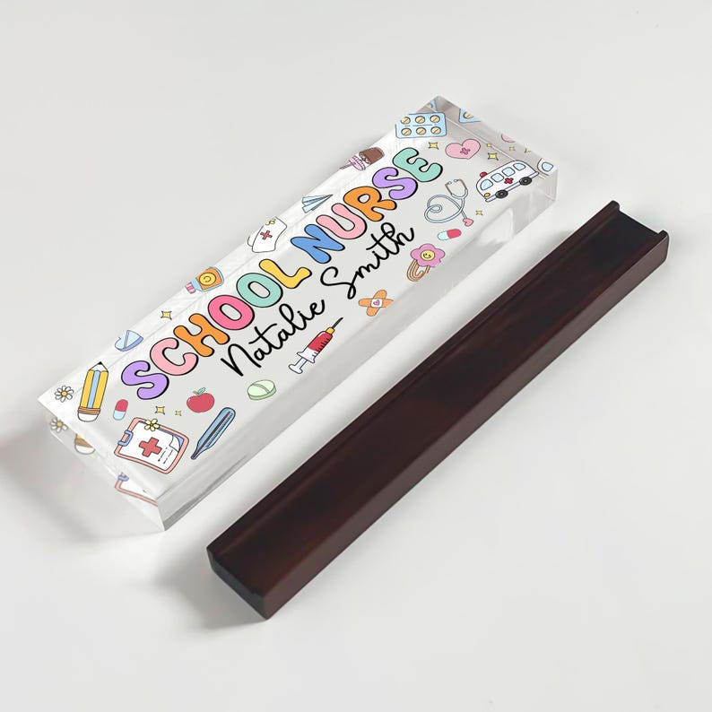 May include: A clear acrylic pen holder with the words "SCHOOL NURSE Natalie Smith" in colorful letters and medical-themed illustrations. Beside it is a dark brown wooden pen holder.