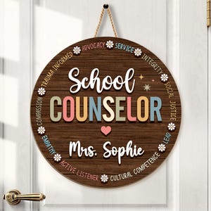 May include: Round wooden sign with the words "School COUNSELOR" in colorful letters. The sign includes the name "Mrs. Sophie" and a heart symbol. The sign is decorated with floral accents and phrases like "Advocacy" and "Integrity".