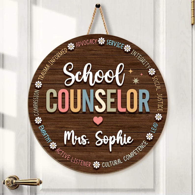 May include: Round wooden sign with the words "School COUNSELOR" in colorful letters. The sign includes the name "Mrs. Sophie" and a heart symbol. The sign is decorated with floral accents and phrases like "Advocacy" and "Integrity".