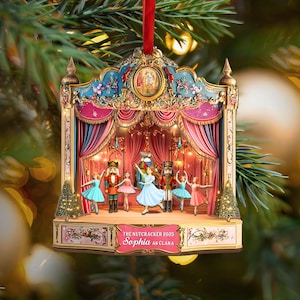May include: A Christmas tree ornament showing a stage from The Nutcracker ballet. The ornament features dancers, a red curtain, and the text "Sophia as Clara". The ornament is trimmed in gold and has a red ribbon for hanging.