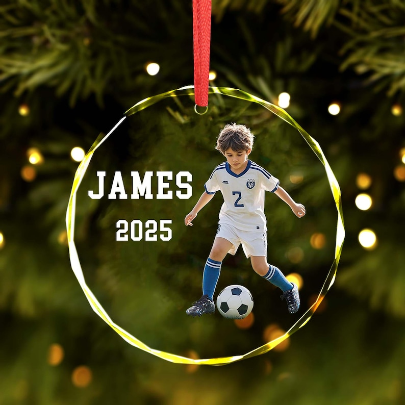 May include: A clear, round Christmas ornament with the name "JAMES" and the year "2025" printed in white. The ornament features a young boy in a soccer uniform kicking a soccer ball. The ornament hangs from a red ribbon.