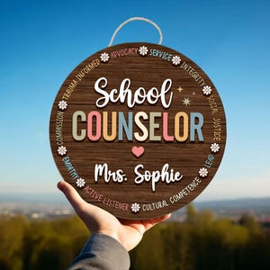 May include: A round wooden sign with the words "School Counselor" in colorful letters. The sign includes the name "Mrs. Sophie" and a heart symbol. The sign has a rope for hanging and is decorated with floral accents and words like "Advocacy" and "Empathy".