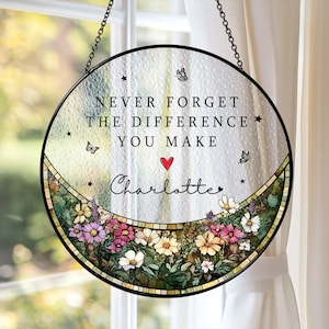 May include: A round, stained-glass window hanging with a black metal frame and chain. The window features the text "NEVER FORGET THE DIFFERENCE YOU MAKE" above the name "Charlotte" and a floral border with butterflies and stars.