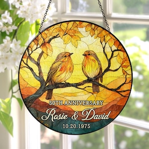 May include: A round stained glass window hanging with a black metal frame and chain. The artwork features two orange and yellow birds perched on a tree branch, with a sunset background. Text reads "50th Anniversary" and "Rosie & David 10.20.1975."