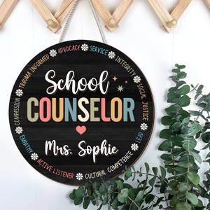 May include: Round wooden sign with the words "School Counselor" in colorful letters. The sign includes the name "Mrs. Sophie" and a heart symbol. The sign is decorated with words like "Advocacy" and "Integrity" around the border.