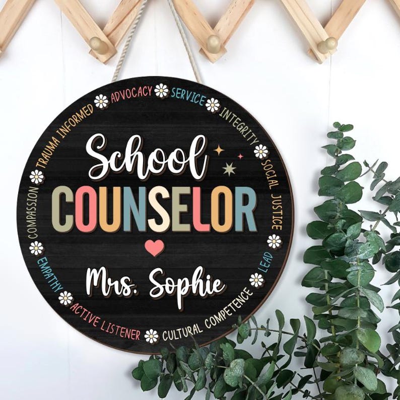 May include: Round wooden sign with the words "School Counselor" in colorful letters. The sign includes the name "Mrs. Sophie" and a heart symbol. The sign is decorated with words like "Advocacy" and "Integrity" around the border.