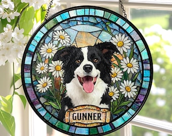 May include: Round stained glass window art featuring a black and white dog with a pink tongue, surrounded by white daisies. The dog's name, "GUNNER," is displayed on a banner. The window is framed in black and hangs from a chain.