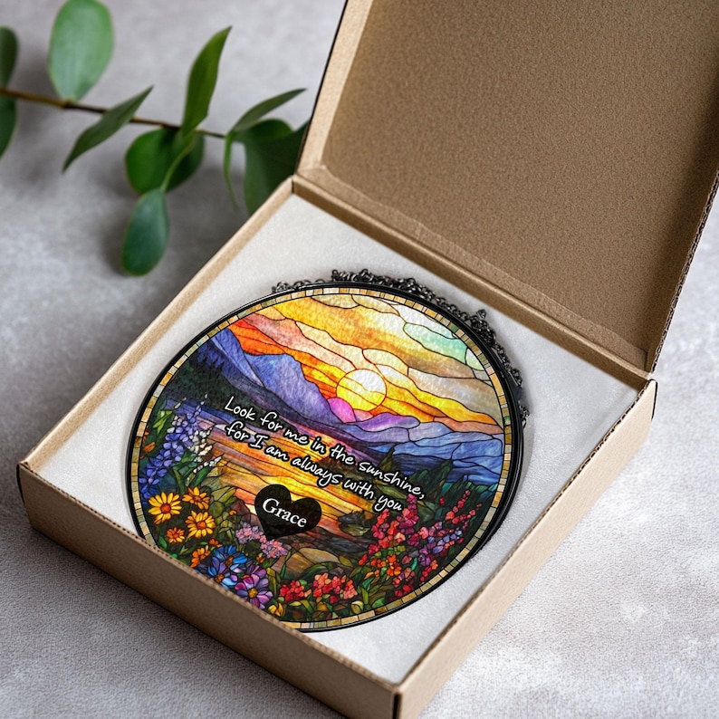 May include: A circular stained glass suncatcher featuring a sunset landscape with mountains, flowers, and a black heart-shaped plaque with the name Grace. The text reads: Look for me in the sunshine, for I am always with you. It is inside a cardboard box.