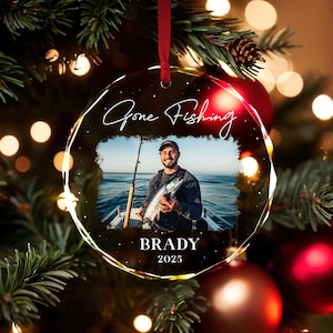 May include: A clear, round Christmas ornament with the words "Gone Fishing" in script. The ornament features a photo of a person holding a fish. The name "Brady" and the year "2025" are below the photo. The ornament hangs from a red ribbon.