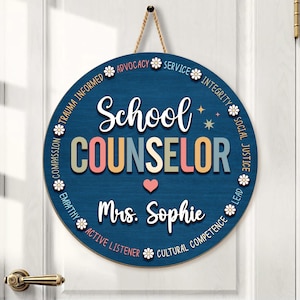 May include: A round, navy blue wooden sign with the words "School COUNSELOR" in colorful letters. The sign includes the name "Mrs. Sophie" and is decorated with floral accents and phrases like "Advocacy" and "Empathy". A jute rope is attached for hanging.