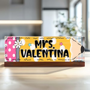 Könnte beinhalten: Ein rechteckiges Schreibtischschild aus Acryl in Stiftform mit gelbem Körper, rosa Radiergummi-Ende und weißen Punkten. Der Text "MRS. VALENTINA" ist in Schwarz gedruckt, ergänzt durch eine weiße Blume sowie kleine blaue und rosa Sterne.