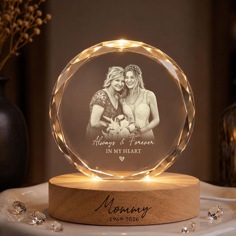 May include: A circular crystal plaque with a faceted edge, illuminated from a light-coloured wooden base. The crystal features an etched photograph of two people and the text "Always & Forever IN MY HEART" with a heart icon. The base displays "Mommy 1969-2026".