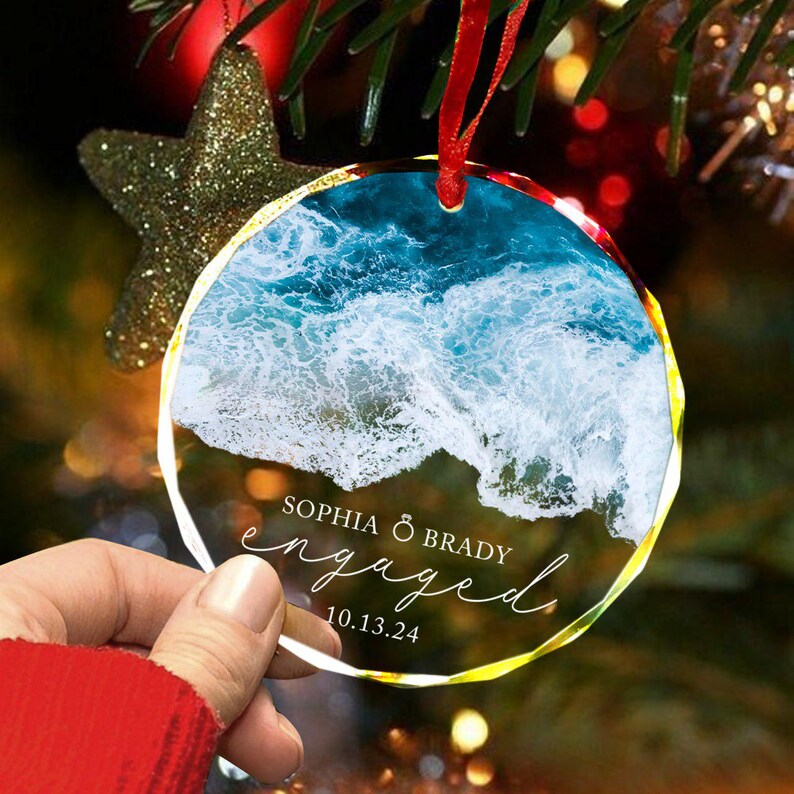 May include: A clear glass ornament with a photo of ocean waves and the text "Sophia & Brady engaged 10.13.24"
