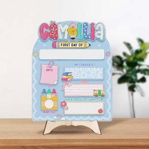 K&ouml;nnte beinhalten: Ein hellblaues Holzschild mit dem Namen "Camellia" in bunten Buchstaben. Das Schild ist f&uuml;r den ersten Schultag gedacht und enth&auml;lt Abschnitte f&uuml;r Datum, Alter, Lehrer und W&uuml;nsche. Es wird von einer Holzstaffelei getragen.