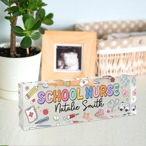 May include: A clear acrylic block with the words "SCHOOL NURSE" in colorful bubble letters and the name "Natalie Smith" in black script. The block is decorated with medical-themed illustrations like a stethoscope, ambulance, and pills.