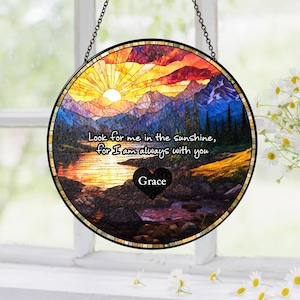 May include: A circular stained glass suncatcher featuring a sunset landscape with mountains, a lake, and trees. The text reads, "Look for me in the sunshine, for I am always with you," above a black heart shape with the name "Grace" printed in white.