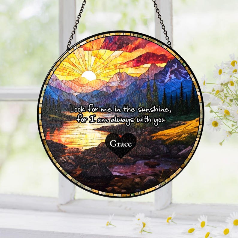 May include: A circular stained glass suncatcher featuring a sunset landscape with mountains, a lake, and trees. The text reads, "Look for me in the sunshine, for I am always with you," above a black heart shape with the name "Grace" printed in white.