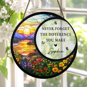 May include: A round, stained-glass window hanging with a black metal frame. The left side features a sunset over a lake and mountains, while the right side has the text "NEVER FORGET THE DIFFERENCE YOU MAKE" with a heart and the name "Sophia".