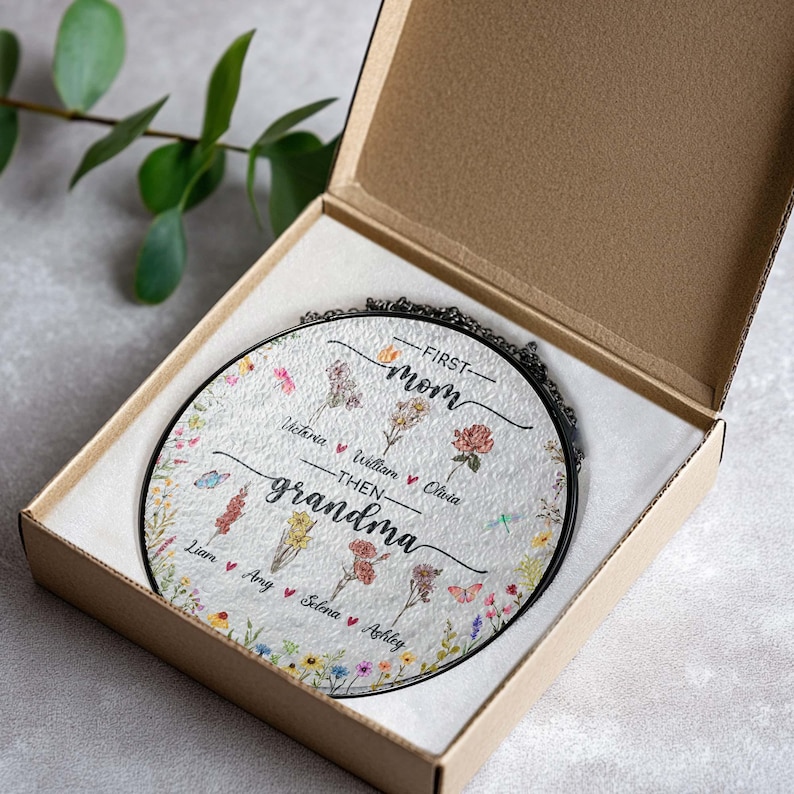 May include: A round glass ornament with a floral design and the text "First Mom Then Grandma" with names listed below. The ornament is in a brown box with a white interior.
