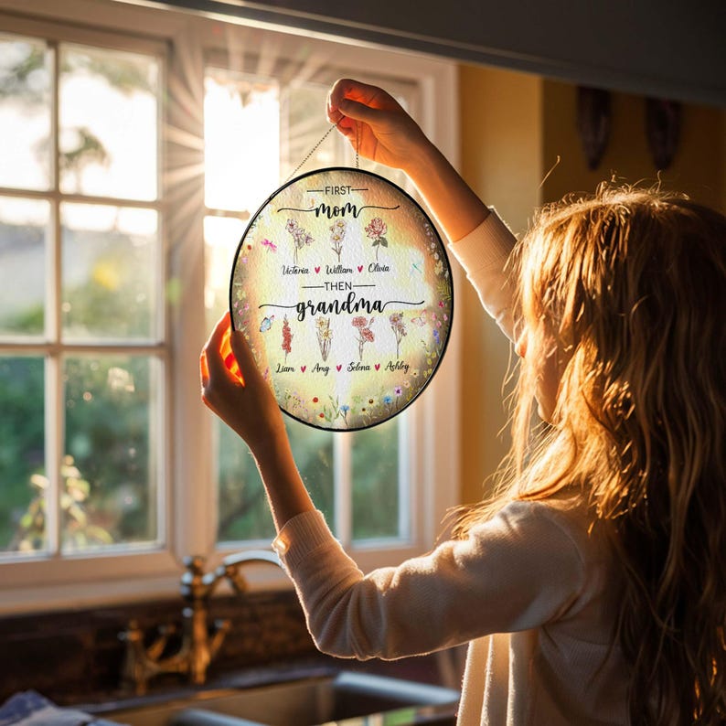 May include: A round, floral-themed sign with the text "First Mom Then Grandma" and a list of names: Victoria, William, Olivia, Liam, Amy, Selena, and Ashley. The sign is hanging from a chain and is being held by a person.