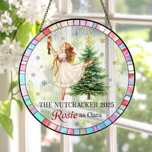 May include: Circular stained glass window depicting a ballerina with a nutcracker and Christmas tree. Text reads "THE NUTCRACKER 2025" and "Rosie as Clara". The border has a colourful, geometric design, and the window hangs from a black chain.