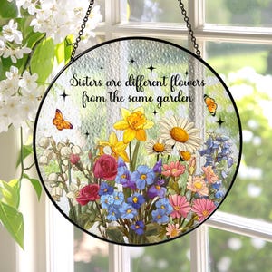 May include: Round stained glass window art with a black metal frame. The artwork features a colorful floral arrangement with butterflies and the text "Sisters are different flowers from the same garden."