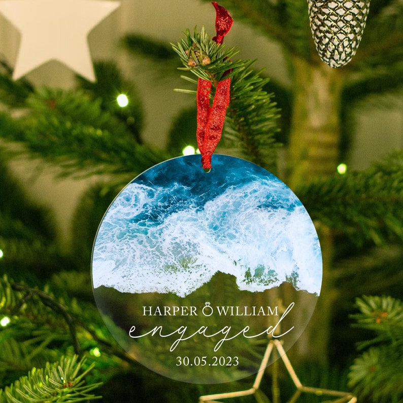 May include: Round clear glass ornament with a photo of ocean waves. The ornament has the text "HARPER & WILLIAM engaged 30.05.2023" printed on it.