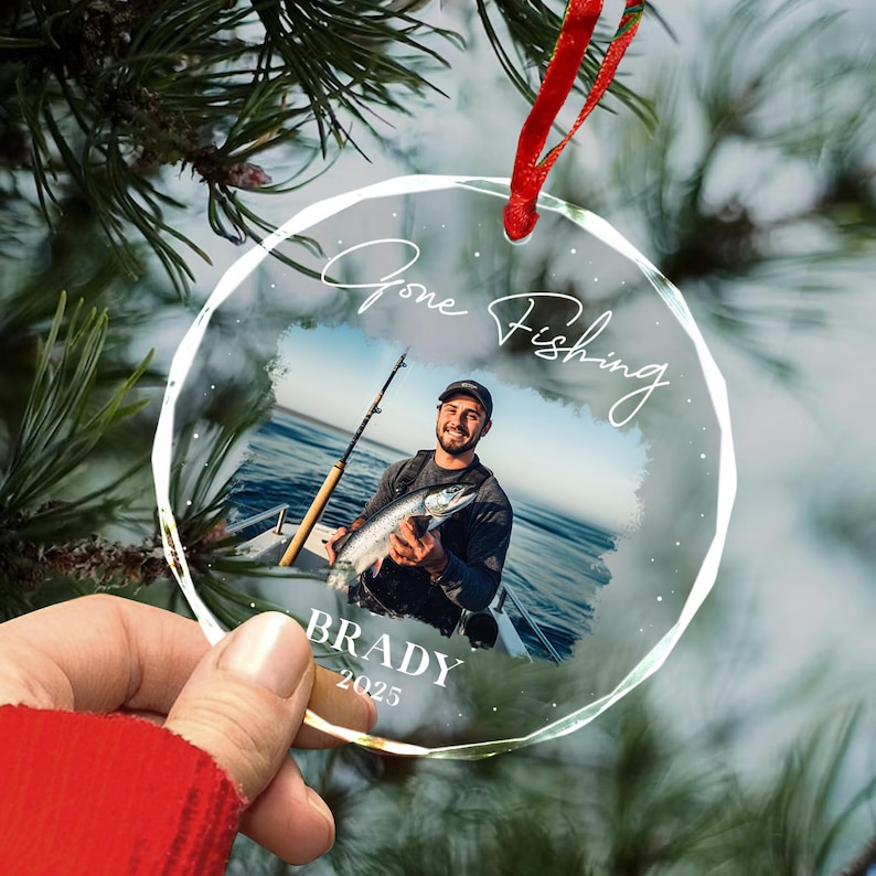 May include: Clear, round Christmas ornament with a photo of a person holding a fish and fishing rod. The ornament has the words "Gone Fishing" and the name "Brady" with the year "2025" printed on it. A red ribbon is attached for hanging.