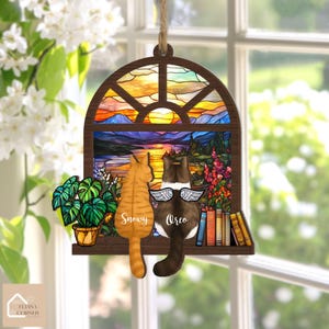 May include: Wooden cat-themed ornament featuring two cats, "Snowy" and "Oreo", gazing out a stained-glass window depicting a sunset over mountains and water. The ornament includes a potted plant and books. The ornament is suspended by a natural twine.
